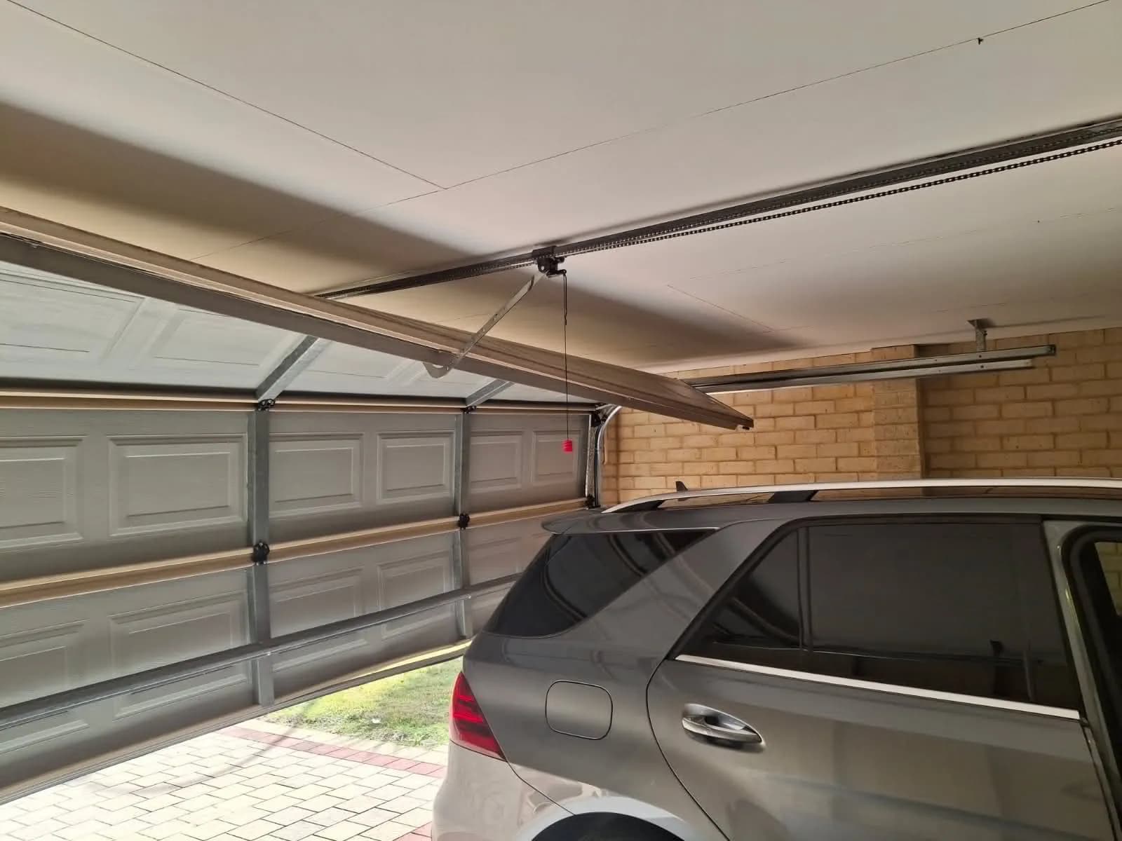 Beige sectional garage door with a bent panel part-open under a carport in Thornlie Beige sectional garage door with a bent panel part-open under a carport in Thornlie